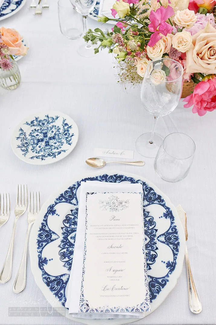 Coperto con piatto maiolicato blu, menu, bicchieri e bouquet di rose rosa e pesca